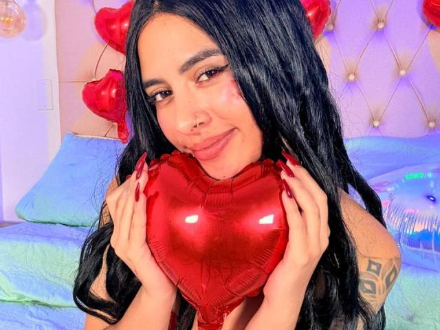 LunaDesire with long black hair, red heart-shaped balloon, and red nails, smiling, holding the balloon to her chest. Tattoo on her right shoulder. Bed with blue pillows.