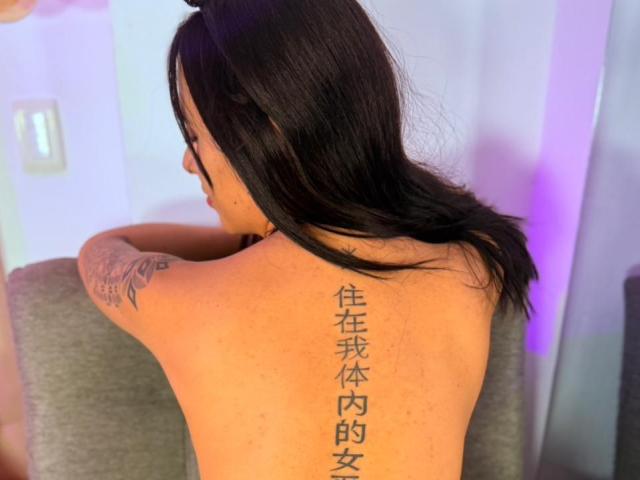 LunaDesire with shoulder-length black hair, back tattoo of Chinese characters, topless, leaning on gray chair, visible arm tattoo, light skin, subtle purple lighting in background.