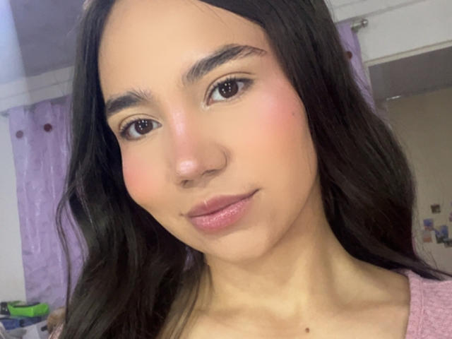 HannaCarlton's face with long black hair, brown eyes, light skin, small nose, pink lips, and subtle makeup, wearing a pink top, smiling softly.
