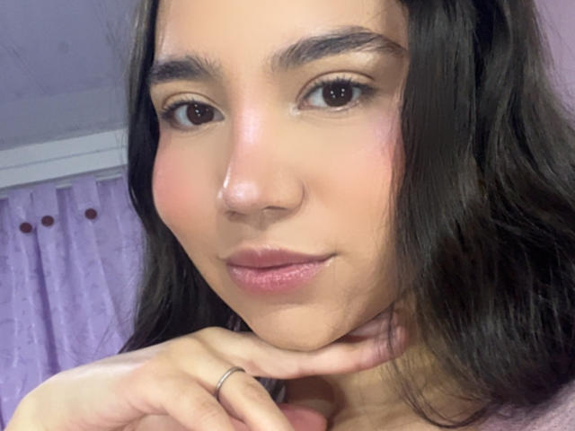 HannaCarlton's face, brown eyes, and pinkish lips, with long dark hair, slight blush, and a ring on her right hand, resting her chin on her hand.