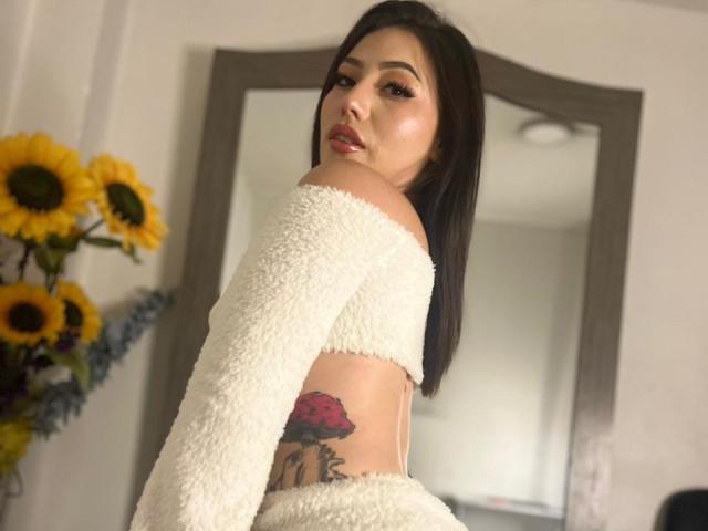 MiaParys with medium brown hair, white fluffy crop top and sleeves, looking over shoulder, showing a colorful rose tattoo on her side, with sunflowers in the background.