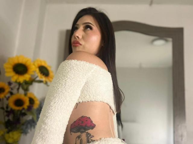 MiaParys with long black hair, white fuzzy off-shoulder top, showing a red mushroom tattoo on her side, looking over her shoulder, sunflowers in the background.