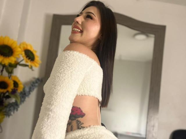 MiaParys with a fluffy white off-shoulder top, smiling, showing a colorful tattoo on her side, and her ass in the background. Sunflowers in the left.