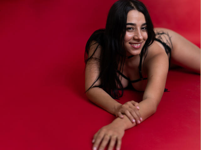 SammyRodriguez is lying on a red surface, smiling, with black hair, wearing black lingerie, showing her tits, and her hands are in front of her, looking cute.