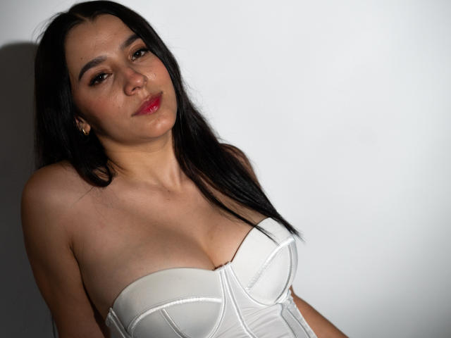 SammyRodriguez with long black hair, red lipstick, and white strapless bustier, showing her tits, looking slightly to the side with a relaxed expression.