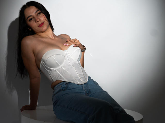 SammyRodriguez with long black hair, white strapless corset, and blue jeans, sitting on white surface, hand on tits, slight smile, shadow on wall, watch on left wrist.