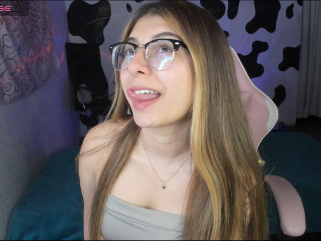 AbbyDew with long brown hair, glasses, and a cute smile, showing her tits in a strapless top, wearing a gold necklace, sitting on a pink chair.