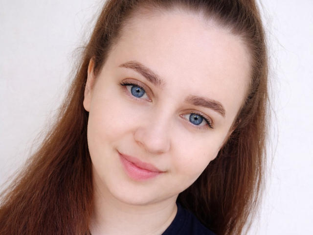 Close-up of KimKistina with light skin, blue eyes, and brown hair in a ponytail. She has subtle eyeliner, thin eyebrows, and a slight smile. Her face is centered, with soft lighting highlighting her smooth skin and minimal makeup. She wears a dark blue shirt. The image focuses on her facial features and expression.