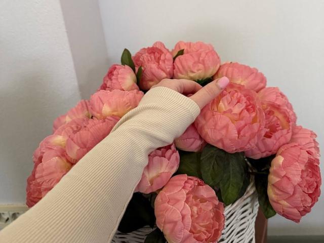 Diantrisy's hand in a ribbed, cream-colored sleeve touches a bouquet of pink peony flowers with green leaves in a white basket.