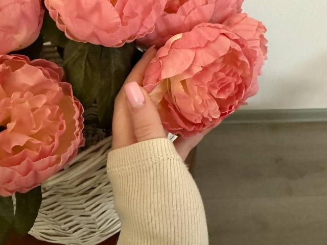 La mano di Diantrisy, con le unghie dipinte di rosa, tiene un cesto di vimini pieno di fiori di peonia rosa, mentre la sua manica beige a coste copre le dita..