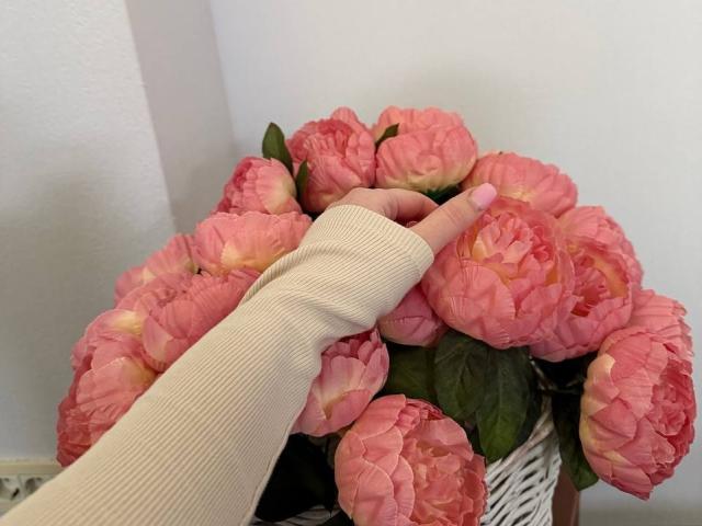 Diantrisy's hand with a pink manicure points at a bouquet of pink peony flowers, her arm in a ribbed, beige long-sleeve shirt.