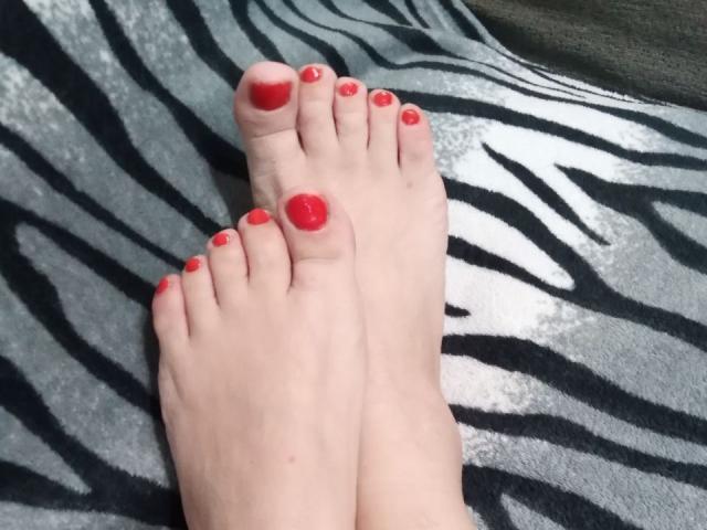 Two pale feet with red-painted toenails lie on a zebra-patterned blanket. The feet are positioned with soles facing up, toes slightly spread. The red nail polish is glossy and evenly applied. The zebra pattern features bold black and white stripes. The texture of the blanket is soft and plush. The feet are centered in the frame, with a slight shadow on the right side. The image is sharp, with clear details of the feet and blanket.