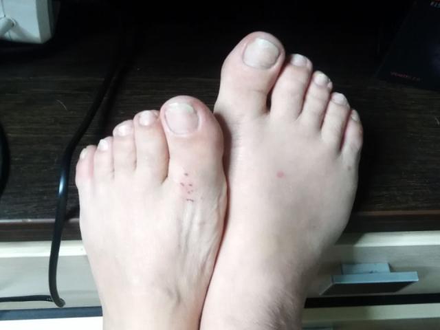 Close-up of IllusionGold's feet on a dark wooden surface. The feet are pale with slightly rough skin and small red blemishes on the left foot's heel. IllusionGold's toenails are short and slightly uneven. The feet are slightly overlapping, with the right foot on top. A black cord is visible on the left side, and a white drawer handle is on the lower right. The image has a natural, unfiltered look with soft lighting.