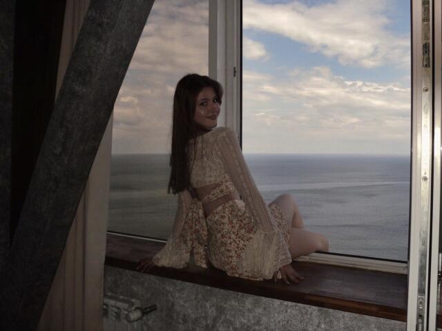 Nikollia mit langen, braunen Haaren, in einem Spitzenkleid, sitzt auf einem Fensterbrett, blickt mit einem Lächeln zurück, ihr Hintern ist leicht angehoben, draußen ist das Meer und ein bewölkter Himmel zu sehen..