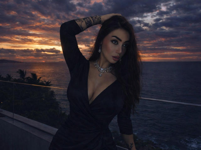 Sophie Lombardo met grote borsten, een donkere jurk, decolleté, tatoeages, hand in haar haar, een ketting, blauwe ogen, donkere lipstick, zonsondergang, oceaan, glazen balustrade, donkere lucht, oranje wolken, sexy..