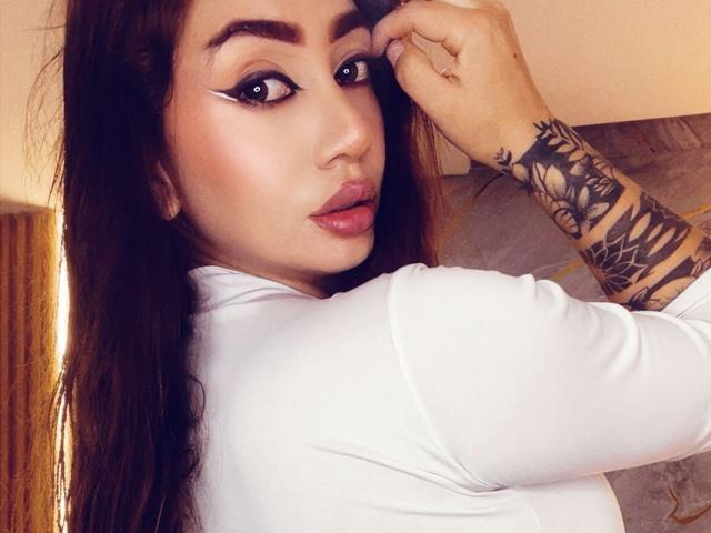 SophieLombardo with long brown hair, white top, black winged eyeliner, pink lipstick, and tattooed arm, looking over shoulder, touching her eyebrow. Her tits are not visible.