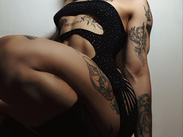 Tattooed YeiSela with polka-dot black dress, showcasing muscular arms and legs, sitting with knees up, displaying various tattoos and a tattooed chest.