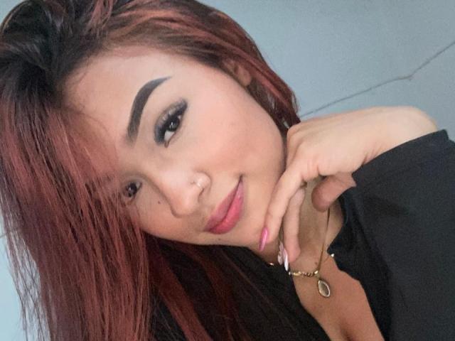 SashaHans with red-tinted hair, black eyebrows, dark eye makeup, pink lipstick, nose ring, black top, hand on chin, necklace, smiling, showing off her cute ass.