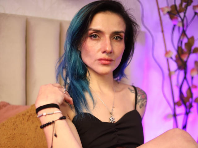 EmaLeBlanc with blue-dyed hair, black tank top, tattoos on right arm, purple-tinged lighting, sitting on bed, looking at camera, black bracelet, nose ring, and pink lipstick.