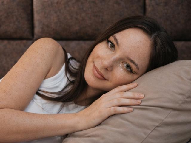 ApheliaAddington, a young woman with fair skin and freckles, lies on a beige pillow, head resting on her hand. She has long, dark brown hair, and her eyes are slightly downcast, giving a subtle, gentle smile. She wears a white sleeveless top. The texture of the brown, padded background contrasts with the softness of the pillow. The image has a relaxed, intimate feel. The subtle, natural makeup enhances her freckles.