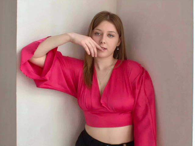 AstiAngel-hot with straight brown hair, wearing a pink satin top, has her hand near her pink tits, looking at the camera with a subtle smile.
