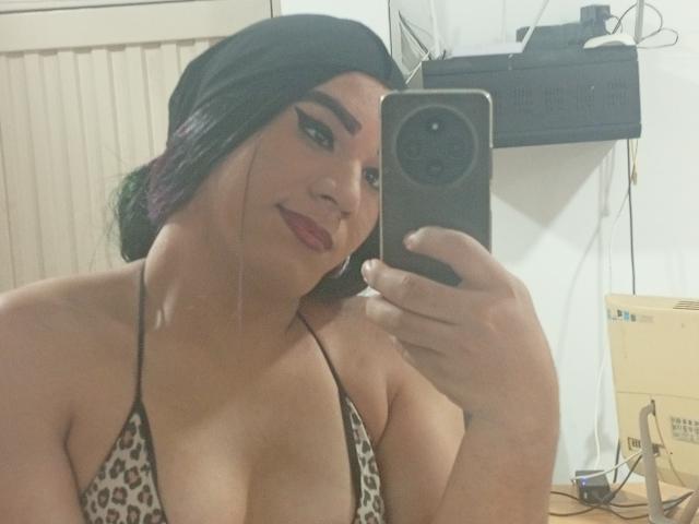 MiaSims con i capelli scuri, che indossa un foulard nero, un top da bikini leopardato che mette in mostra un seno prosperoso, tiene un telefono, sorride, sta scattando un selfie allo specchio, in una stanza..