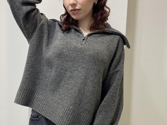 A photo of RuthanneWelliver. She has wavy, dark red hair and is wearing a loose, gray, zip-up sweater with a wide, ribbed bottom. Her right arm is raised, and her lips are slightly pursed. The sweater's texture is soft and slightly fuzzy. She is also wearing dark jeans. The background is a plain, off-white wall. The image is focused on the upper half of RuthanneWelliver's body.