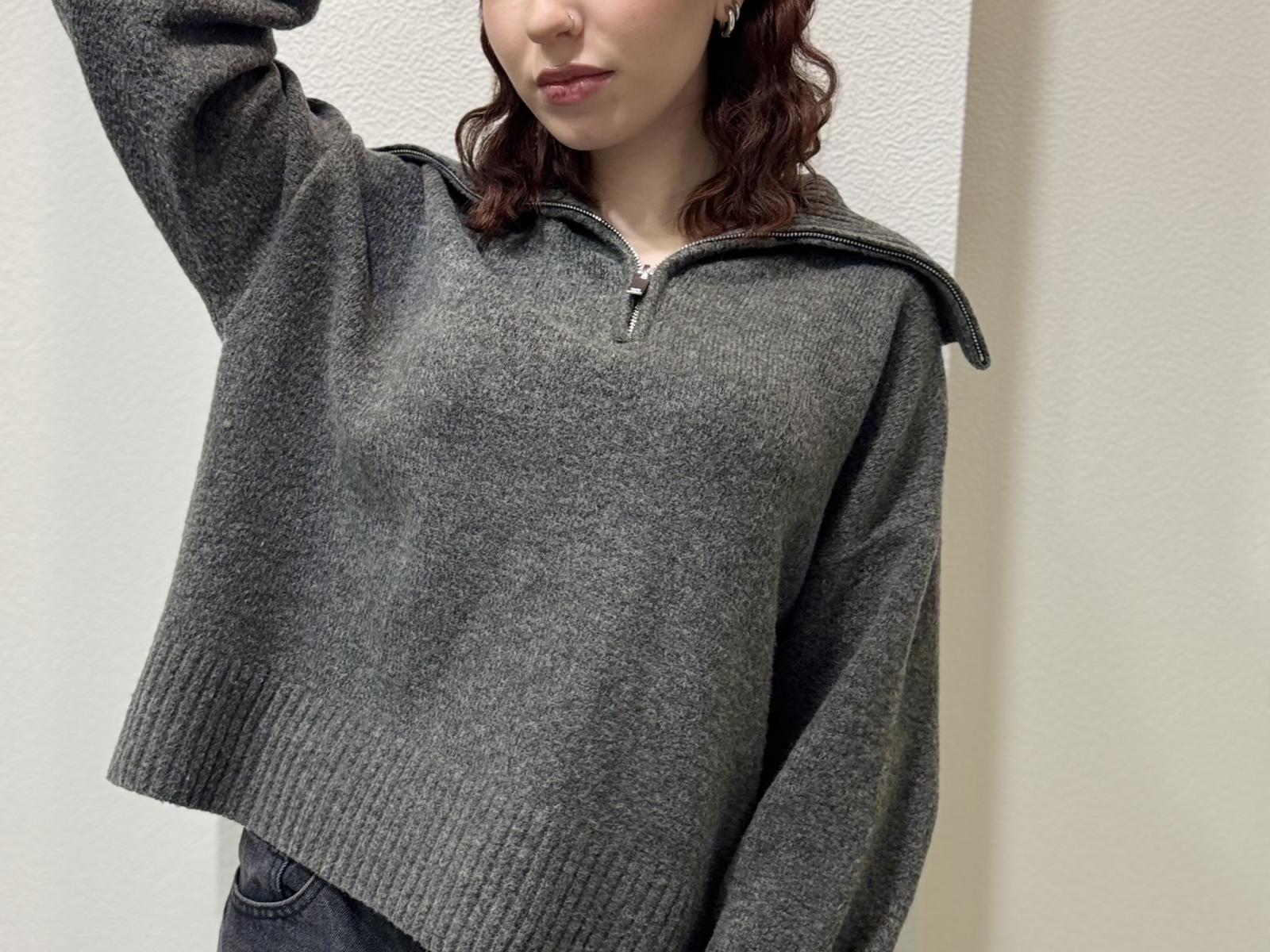 RuthanneWelliver with wavy red hair, wearing a grey, zip-up, oversized sweater, her arm raised, showing a small portion of her tits, standing against a white wall.