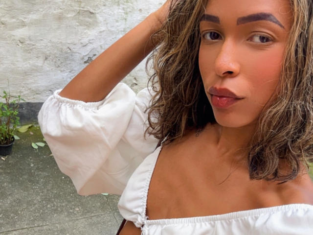 Priscilinda has wavy brown hair, tan skin, and is wearing a white off-shoulder top, showing her small tits and pinkish lips with a relaxed, confident expression.