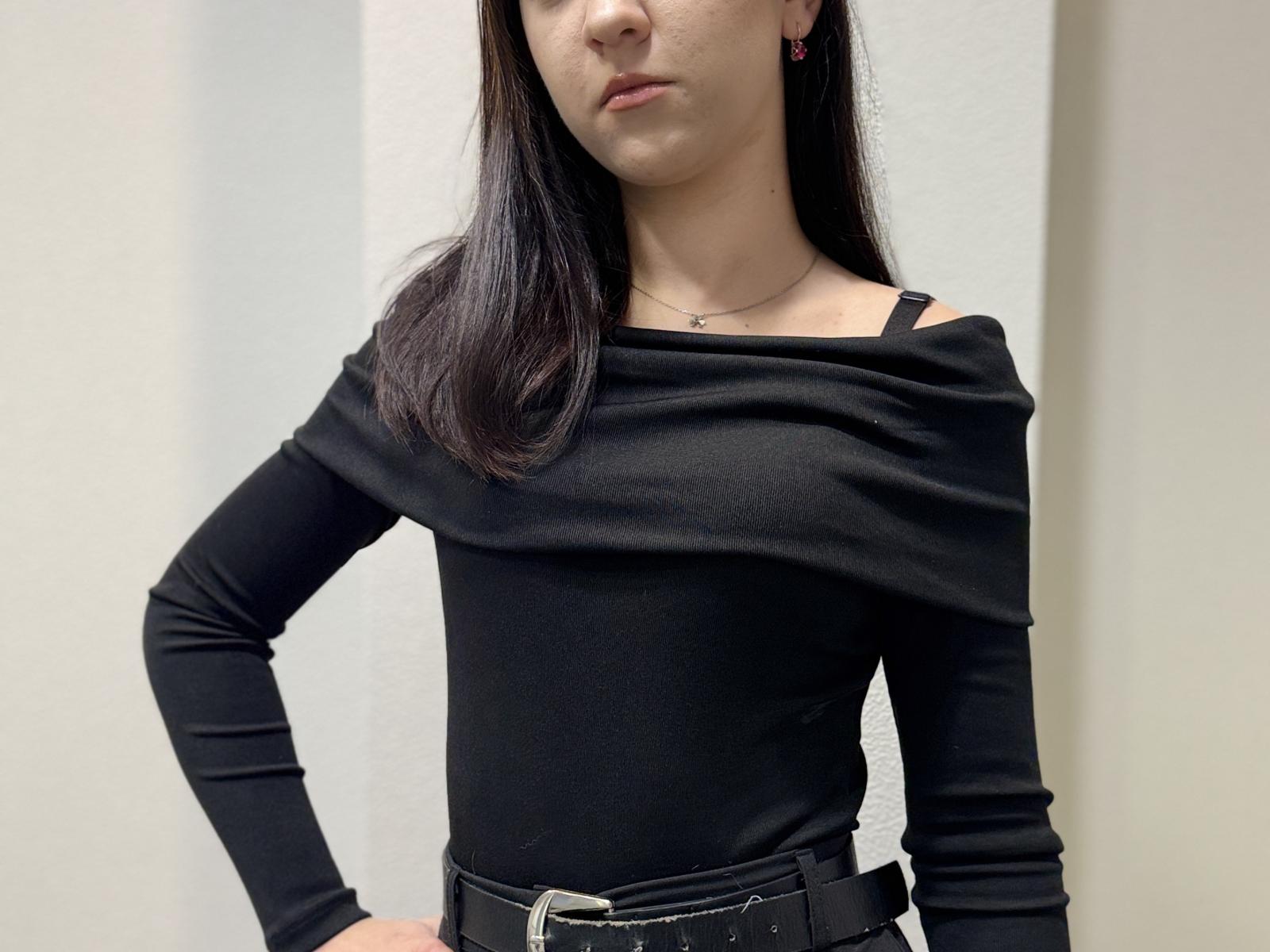 RandyHeyen with dark straight hair, wearing a black off-shoulder top, black belt, and a thin necklace, standing with hands on hips, looking slightly down.