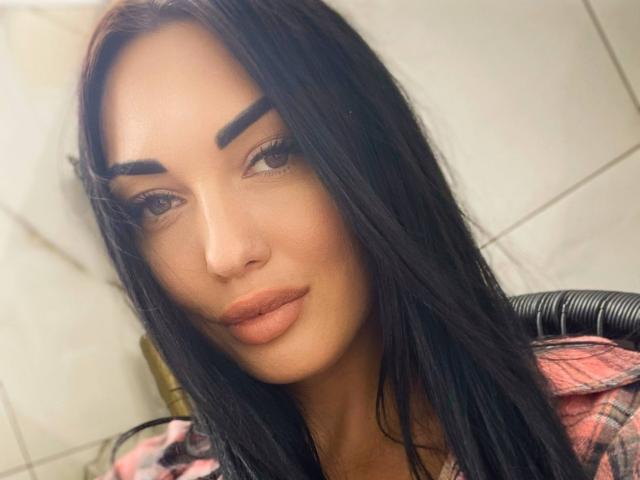 Xioni has long black hair, thick eyebrows, and full orange lipstick. She's wearing a pink plaid shirt, looking directly at the camera with a slight smile.
