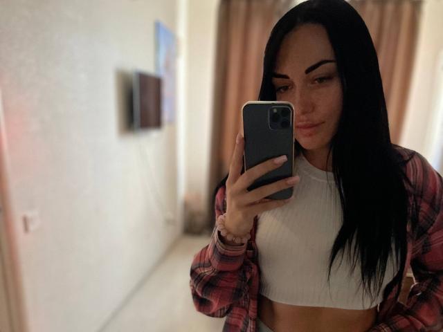 Xioni with long black hair, wearing a red plaid shirt over a white ribbed crop top, taking a mirror selfie with a smirking face, holding a gray phone.
