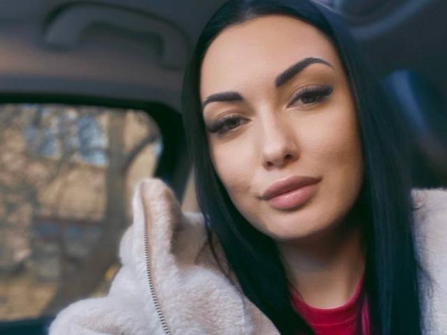 Xioni has a cute face with long black hair, thick eyebrows, and is wearing a fluffy white jacket with a red shirt underneath, looking slightly down.