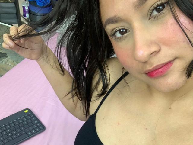 MylaNova with wavy black hair, light skin, red lipstick, black bra, holding hair, slight smile, pink bed, black keyboard in background. Boobs visible, medium size.