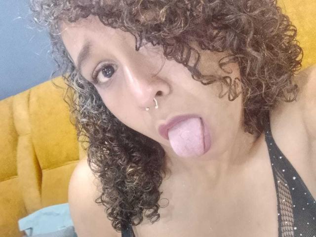 EvelynMonrou with curly brown hair, nose ring, and pink tongue out, wearing a black mesh top, showing her boobs. Yellow headboard in background.