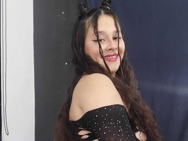 LauretMorras met lang, golvend zwart haar in twee knotjes, lachend met roze lippenstift, gekleed in een zwarte, mouwloze top met glitters, die haar schouders en bovenrug onthult..
