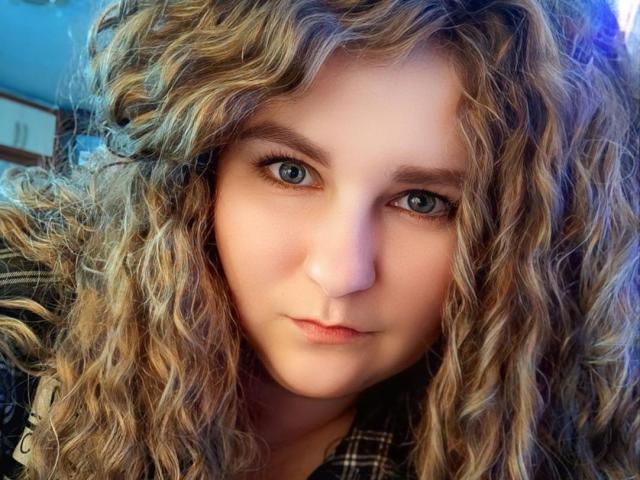 MinistraX has curly blonde hair, blue eyes, and a serious expression. Her nose is small and her lips are slightly closed. She's wearing a plaid shirt.