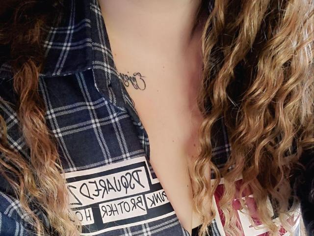 MinistraX's neck with curly brown hair, wearing a blue plaid shirt, showing a small black tattoo just below the jawline. Boob outline visible under the shirt.