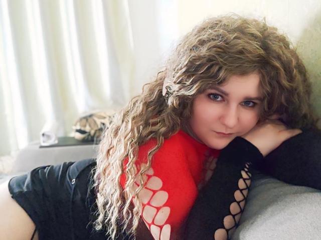MinistraX with curly brown hair, red and black cutout top, and black shorts, lying on a grey couch, head resting on her hand, looking at the camera.
