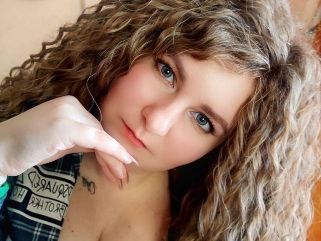 MinistraX has curly brown hair, blue eyes, and a slight blush. She's looking at the camera with a relaxed hand near her face, showing a small chest tattoo.