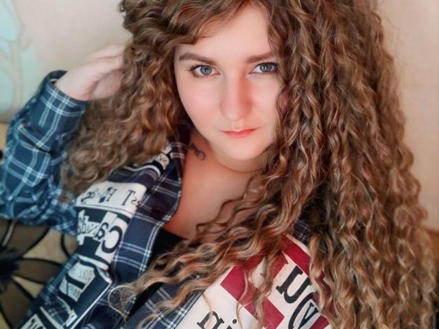 MinistraX with long curly brown hair, blue eyes, and a plaid shirt, looking at the camera with a slight smile, showing a tattoo on their chest, and a cute face.