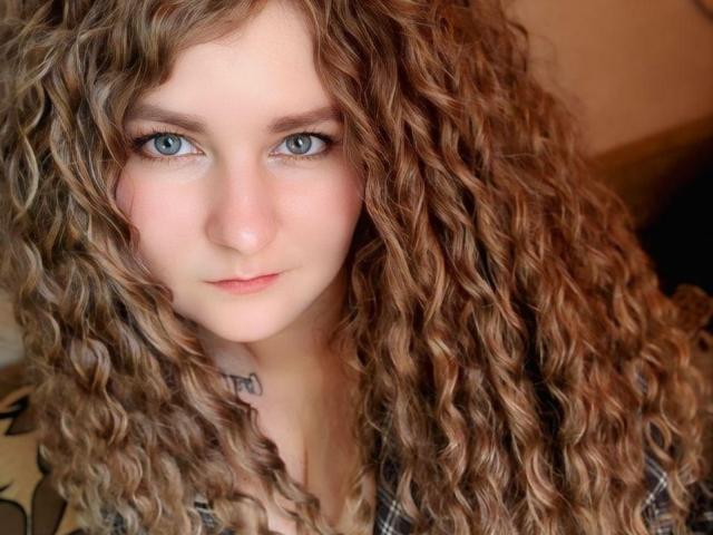 MinistraX has curly brown hair, blue eyes, and a tattoo on her neck. She's looking directly at the camera with a slight, serious expression. Her tits are small and cute.