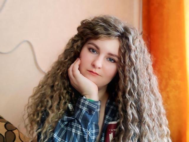 MinistraX with long, curly brown hair, blue eyes, and a hand on her cheek, wearing a blue plaid shirt, looking thoughtful. She's got a cute, soft ass.