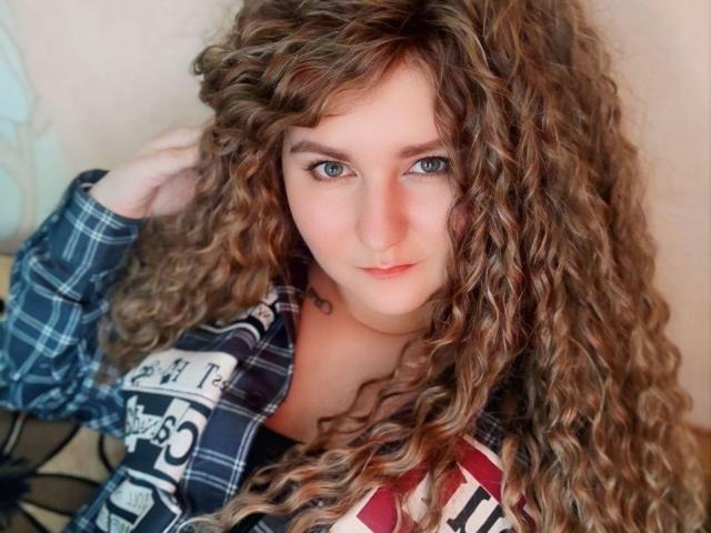 MinistraX with curly brown hair, blue eyes, and a plaid shirt, looking up with a slight smile. Her tits are partially visible under the shirt.