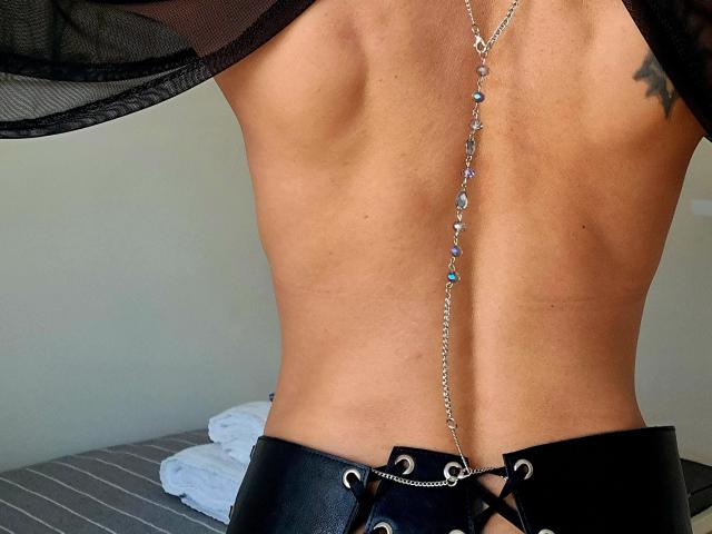 Close-up of DreamityCamy's torso, showing a toned, muscular back with a silver chain necklace hanging down. The necklace has blue and clear beads, leading from the upper back to the black leather waistband with silver buckles. DreamityCamy's skin is tan, and a small tattoo is visible on the right side. The black sheer fabric on the upper left is slightly visible. The waistband is tightly fitted, emphasizing the lower back. The bed with a gray striped cover and white pillows is in the background.
