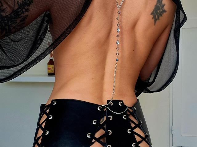 DreamityCamy è tornata con un corsetto nero con lacci, pelle abbronzata in evidenza, un tatuaggio sulla scapola destra, una collana con catena d'argento e un tessuto nero trasparente, che mette in mostra un sedere sexy e tonico..