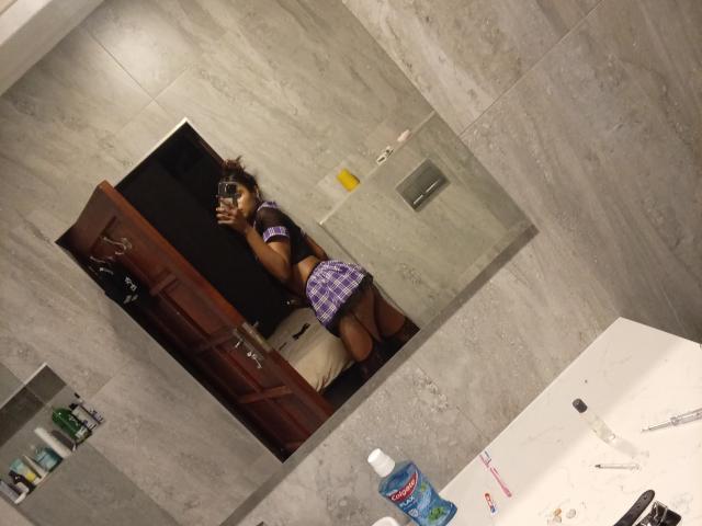 CloeCurlys maakt een selfie in de spiegel, gekleed in een paars geruit topje en een korte rok, waarbij haar donkerbruine achterkant zichtbaar is. Ze houdt een camera vast, en de foto is genomen in een badkamer met grijze tegels..