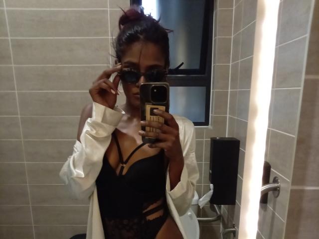 CloeCurlys prend un selfie devant un miroir dans une salle de bain, vêtue d'une chemise blanche, d'un ensemble de lingerie noire à bretelles, et portant des lunettes noires, avec un chignon rouge et tenant un téléphone..