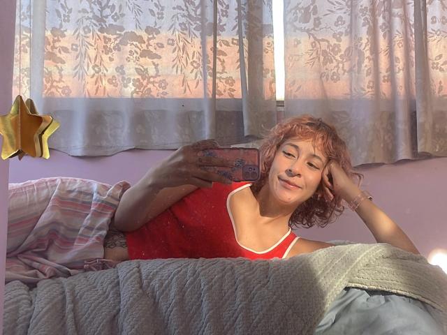 KittyEla met krullend roze haar, een rode tanktop, liggend in bed, een telefoon vasthoudend, lachend, zonlicht op haar gezicht, een geborduurde deken, een gestreept kussen, een gele ster als wanddecoratie, ontspannen, schattig..