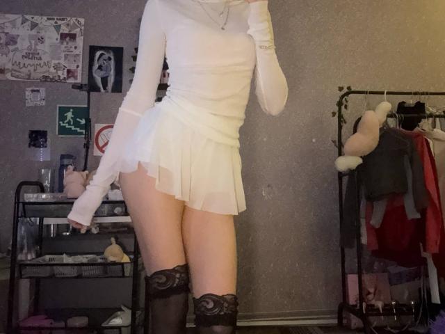 AmilySunray in a white, long-sleeve, sheer top, and a short, white, pleated skirt, with black lace thigh-highs, standing with her hand near her mouth.